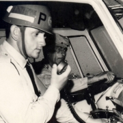 Dois policiais em um carro. Um com um rádio, vestindo um capacete e o outro, na direção, usando uma boina.