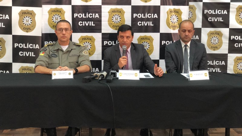 Três homens sentados lado a lado atrás de uma mesa em frente a uma parede com vários Brasões da Polícia Civil. O homem sentado no meio está falando no microfone enquanto olha para frente.