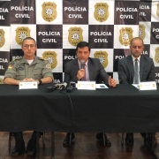 Três homens sentados lado a lado atrás de uma mesa em frente a uma parede com vários Brasões da Polícia Civil. O homem sentado no meio está falando no microfone enquanto olha para frente.