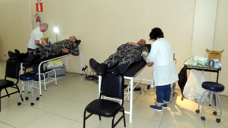 Dois militares estão deitados em macas, dentro de uma sala hospitalar branca, enquanto profissionais da saúde fazem a coleta de sangue.