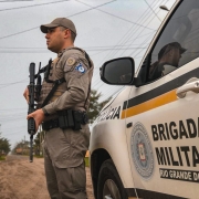 Foto mostra um policial militar de perfil segurando um fuzil ao lado de uma viatura