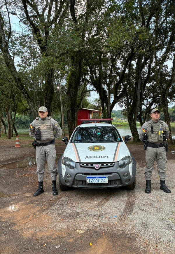 Dupla de PMs e viatura aparecem em uma área arborizada e com um lago ao fundo. 