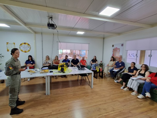Professores reunidos em uma sala, assistindo a fala de uma PM feminina, durante a atividade de recepção, formação e confraternização na Unidade em Passo Fundo. 