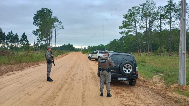 Dois PMs aparecem durante a fiscaliza&ccedil;&atilde;o de um ve&iacute;culo em uma estrada de ch&atilde;o no interior do RS. Ao fundo v&ecirc;-se uma viatura da BM. 