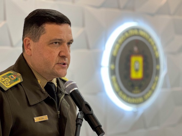 A imagem mostra o comandante-geral da Brigada Militar, coronel PM Luigi Gustavo Soares Pereira, durante pronunciamento em uma solenidade. Ele veste uniforme institucional da Brigada Militar e fala ao microfone. Ao fundo, desfocado, aparece o brasão da Corporação iluminado na parede do auditório. O coronel está voltado para o público enquanto realiza seu discurso.