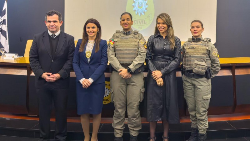 Imagem mostra cinco pessoas posando em um audit&oacute;rio, em frente a uma mesa e a uma tela com o bras&atilde;o da Pol&iacute;cia Civil projetado ao fundo. Duas policiais militares aparecem fardadas, ao centro e &agrave; direita da imagem, e tr&ecirc;s pessoas est&atilde;o em trajes civis. Todos est&atilde;o em p&eacute;, lado a lado, voltados para a c&acirc;mera.