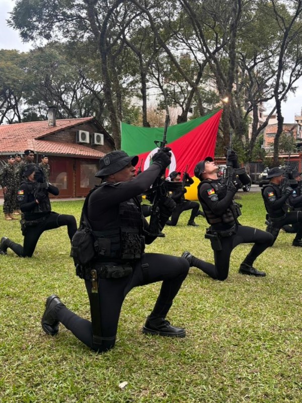 Foto mostra policiais militares ajoelhados em um gramado, com armas apontadas para cima, com a bandeira do RS em fundo