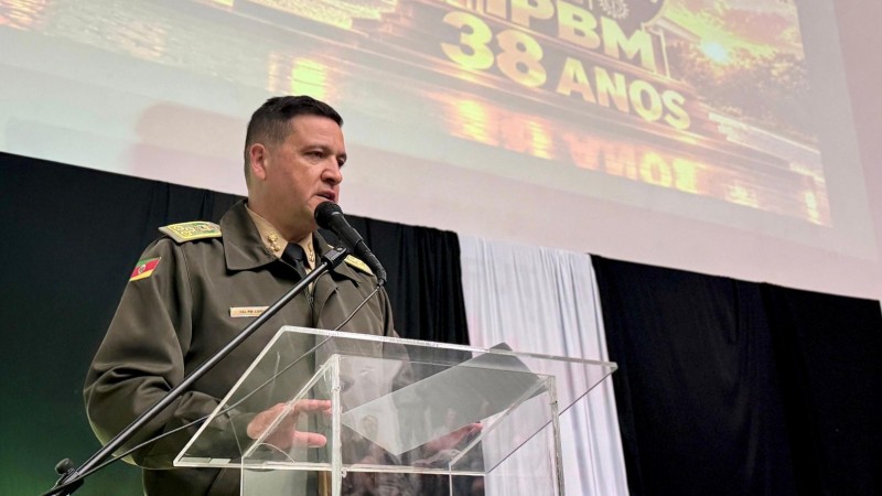 O Comandante aparece diante do p&uacute;lpito falando aos participantes do evento pelo microfone. Veste fardamento e ao fundo h&aacute; um slide e um painel preto com um destaque branco. 