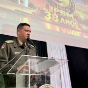 O Comandante aparece diante do p&uacute;lpito falando aos participantes do evento pelo microfone. Veste fardamento e ao fundo h&aacute; um slide e um painel preto com um destaque branco. 